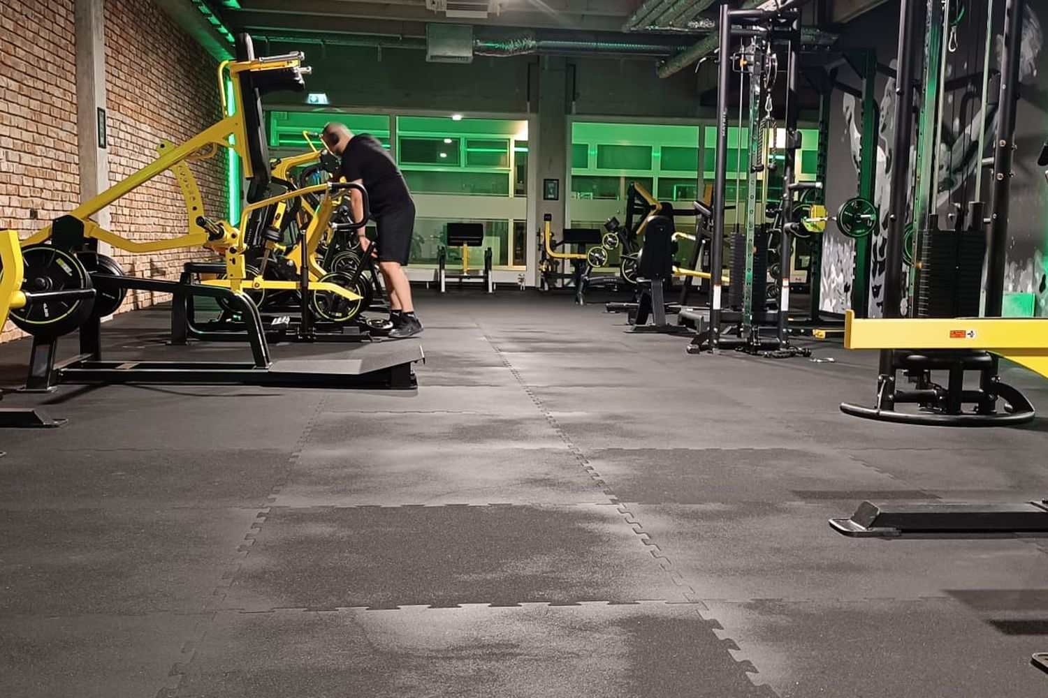 Club floor under green lighting with plate-loaded machines and racks, a technician in the background