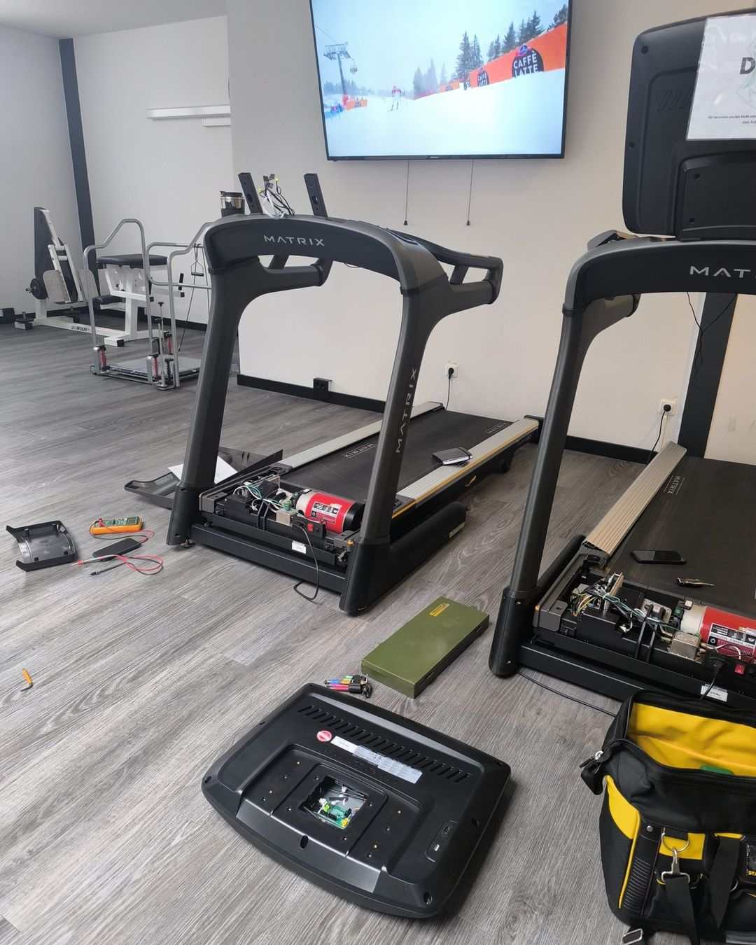 Deux tapis de course avec capot moteur ouvert pendant l’entretien, multimètre et outillage posés au sol