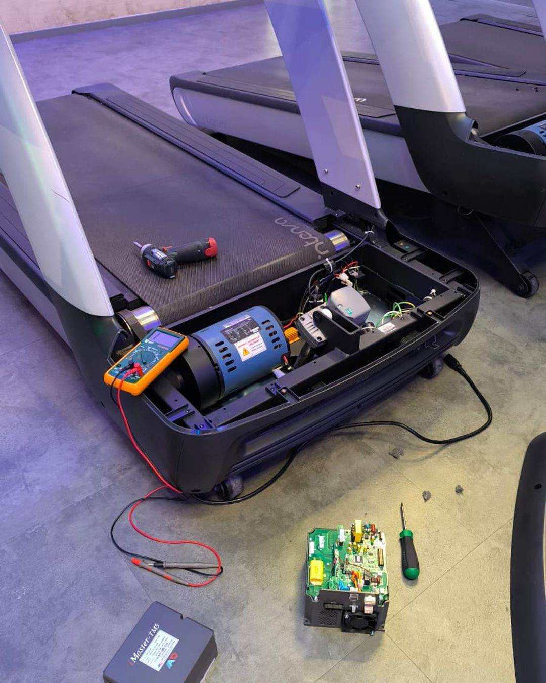 Diagnostic électronique sur un tapis de course : capot moteur ouvert, multimètre sur le moteur, carte de commande déposée au sol