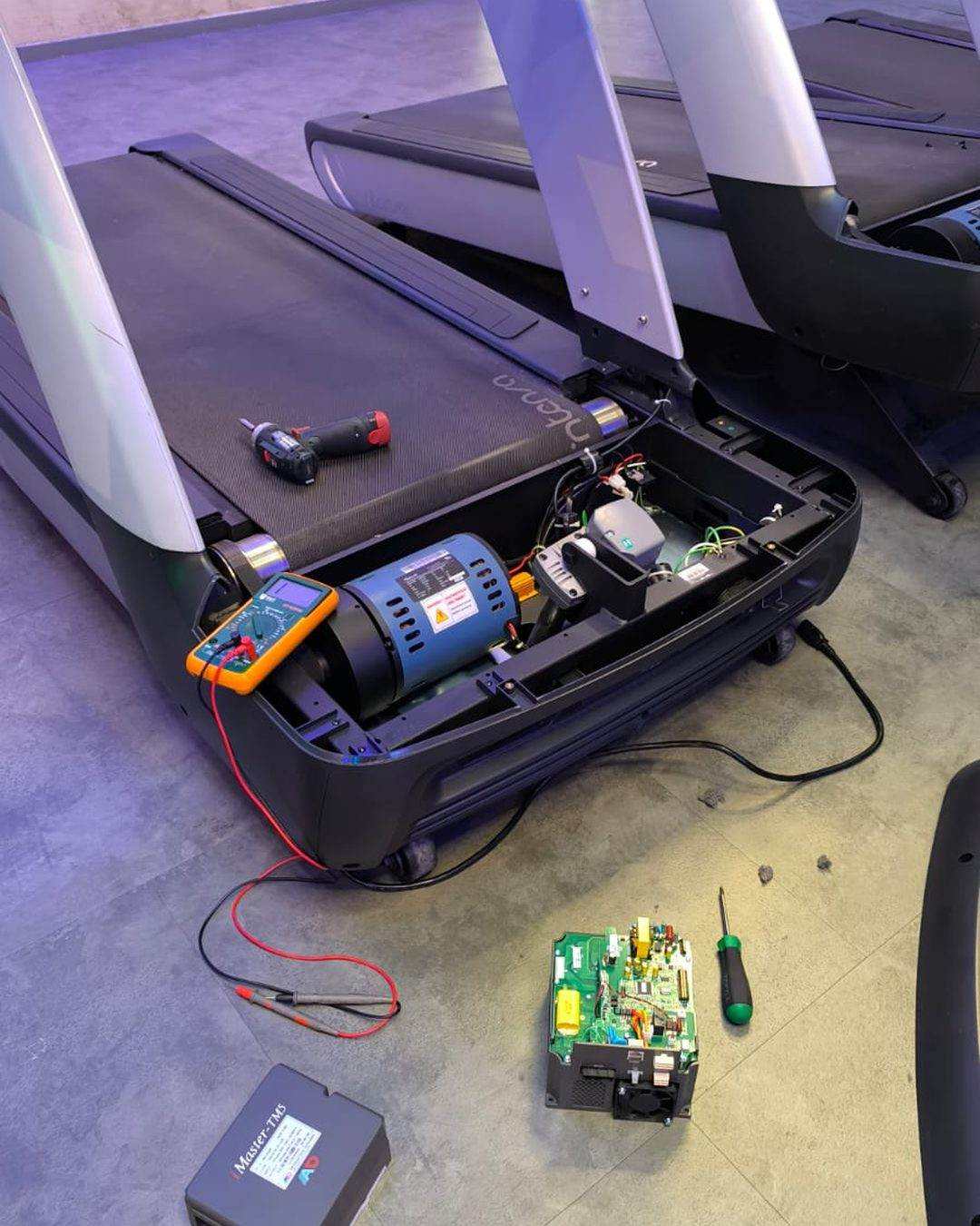 Elektronikdiagnose an einem Laufband: geöffnete Motorhaube, Multimeter am Motor, ausgebaute Steuerplatine am Boden