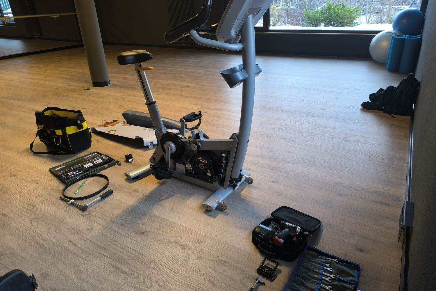 Vélo d’appartement ouvert et outillage déployé au sol avant l’intervention : sacoche, coffret de douilles, clé dynamométrique et courroie