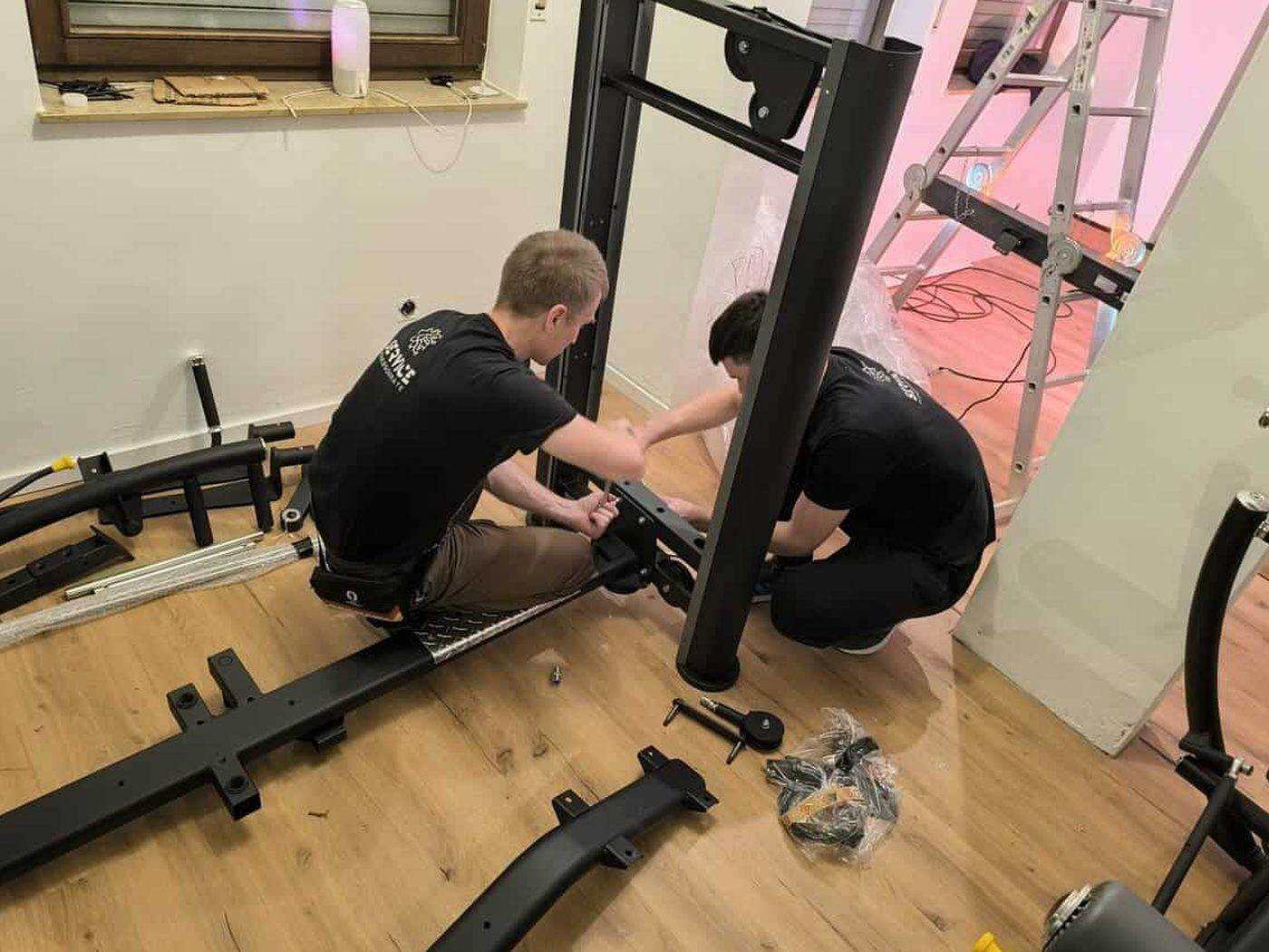 Two technicians assembling a strength station in a private training room, frame parts and bolts laid out on the floor