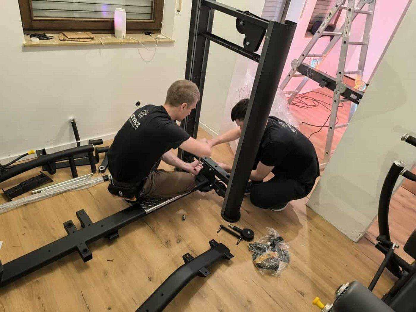 Deux techniciens assemblent une station de musculation dans une salle privée, éléments du châssis et visserie au sol