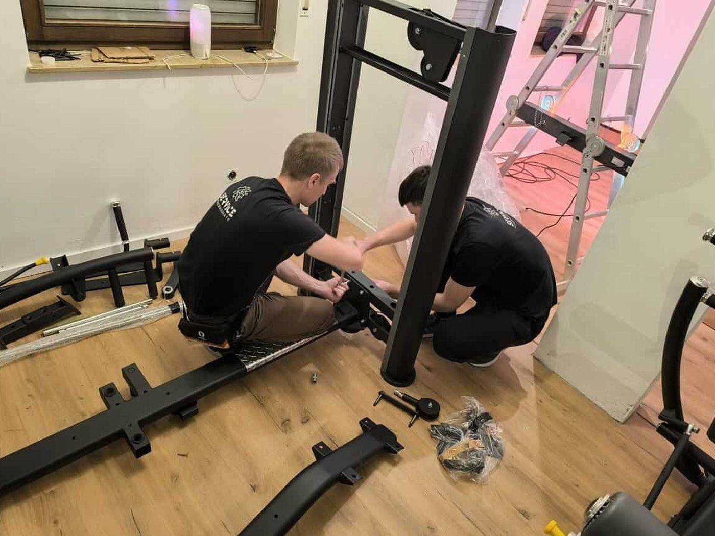 Zwee Techniker bauen eng Kraaftstatioun an engem private Trainingsraum op, Rahmendeeler a Schrauwen um Buedem