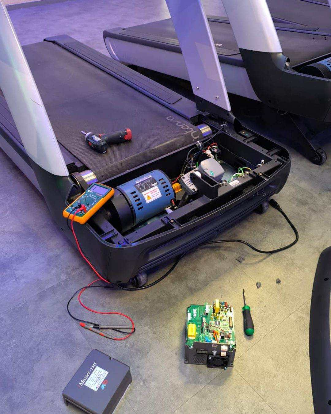 Electronic diagnostics on a treadmill: motor cover open, multimeter on the motor, control board removed and lying on the floor