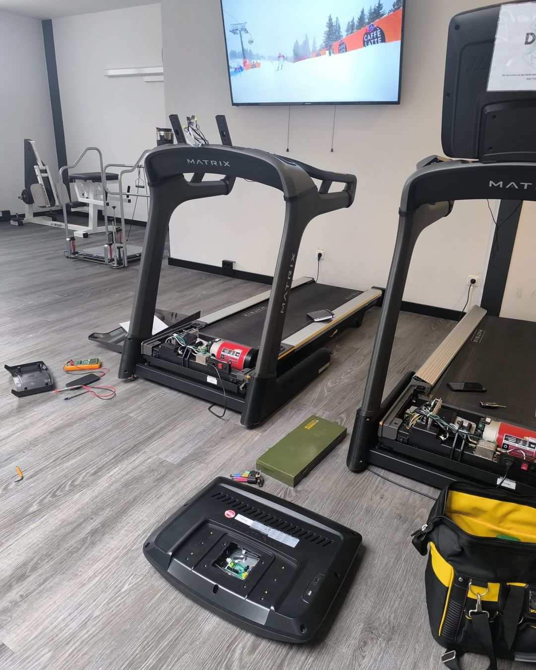 Two treadmills with the motor cover open during servicing, a multimeter and tools on the floor