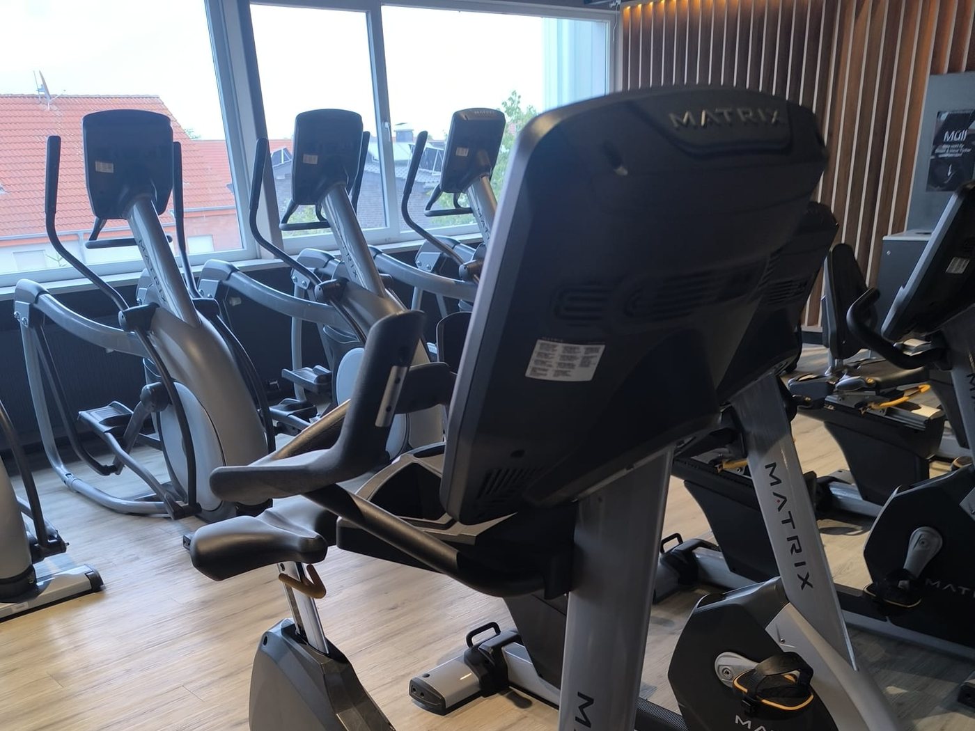 Zone cardio d’une salle de sport avec des vélos d’appartement au premier plan et une rangée de vélos elliptiques devant la baie vitrée